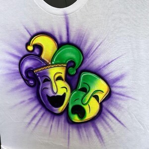 May include: White crew neck t-shirt with an airbrushed design of a jester's hat and two theatrical masks. The masks are yellow, green, and purple, set against a purple burst. The shirt is white.