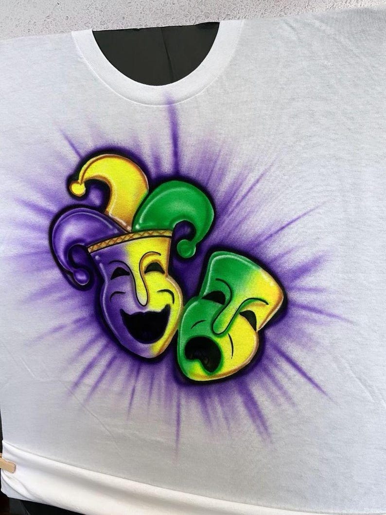 May include: White crew neck t-shirt with an airbrushed design of a jester's hat and two theatrical masks. The masks are yellow, green, and purple, set against a purple burst. The shirt is white.