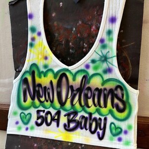 May include: White ribbed vest top with a scoop neckline. The front displays airbrushed text reading "New Orleans 504 Baby" in green, yellow, and purple. Green hearts and purple dots decorate the design. The vest top is set against a dark backdrop.