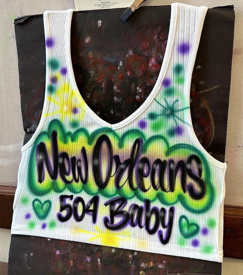 May include: White ribbed vest top with a scoop neckline. The front displays airbrushed text reading "New Orleans 504 Baby" in green, yellow, and purple. Green hearts and purple dots decorate the design. The vest top is set against a dark backdrop.