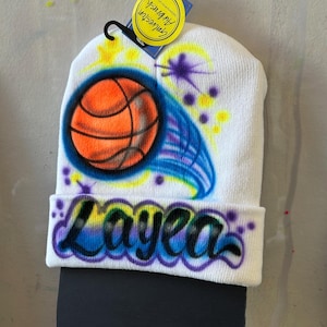 May include: White beanie with a basketball and a blue and purple swirl design. The name "Layea" is written in a colourful font on the beanie.