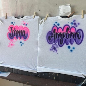 May include: Two white t-shirts with airbrushed names. One shirt says "Jiana" in purple and pink, the other says "Angelina" in purple and pink. Both shirts have decorative starburst and dot designs.