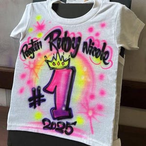 May include: White t-shirt with airbrushed design. Features the names Payton, Ruby, and Nicole in black script, a pink and yellow crown, and the number 1 in pink and purple. Includes the year 2025.