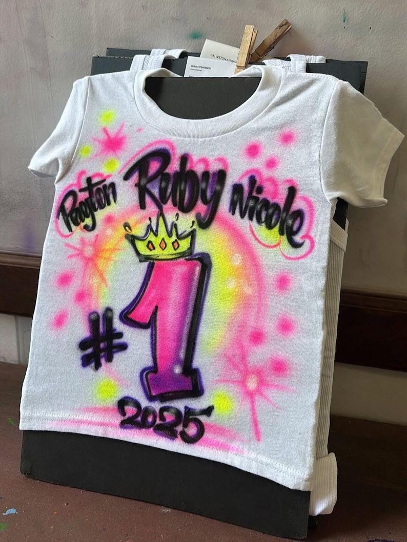 May include: White t-shirt with airbrushed design. Features the names Payton, Ruby, and Nicole in black script, a pink and yellow crown, and the number 1 in pink and purple. Includes the year 2025.