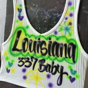 May include: White ribbed vest top with a scoop neck, featuring airbrushed text that reads "Louisiana 337 Baby" in black and green. The design includes yellow starbursts and purple hearts.