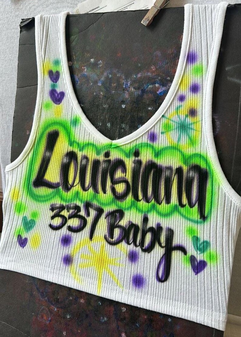 May include: White ribbed vest top with a scoop neck, featuring airbrushed text that reads "Louisiana 337 Baby" in black and green. The design includes yellow starbursts and purple hearts.