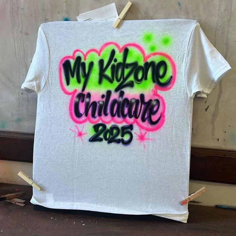 May include: White t-shirt with airbrushed text that reads "My Kidzone Childcare 2025". The text is in black, outlined in pink, and set against a green cloud-like background. Pink starbursts are at the bottom.