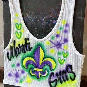 May include: White ribbed vest top with a V-neck, featuring airbrushed text and graphics. The design includes a purple and yellow fleur-de-lis, the words "Mardi Gras", and green and yellow accents.