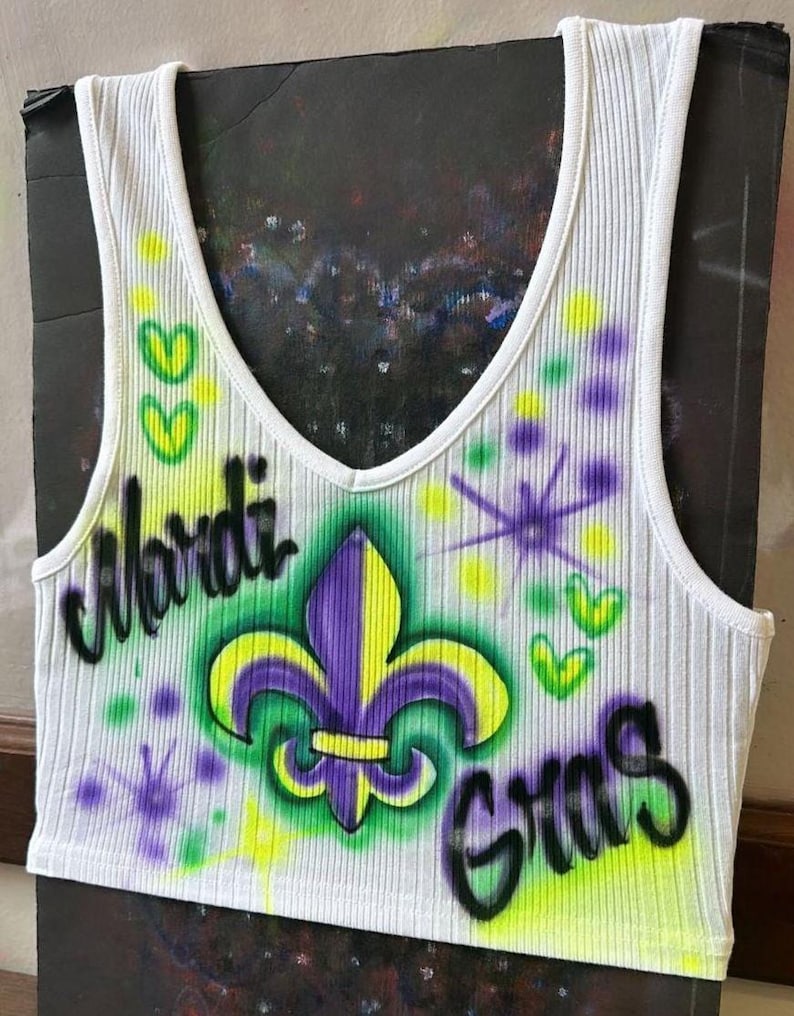 May include: White ribbed vest top with a V-neck, featuring airbrushed text and graphics. The design includes a purple and yellow fleur-de-lis, the words "Mardi Gras", and green and yellow accents.