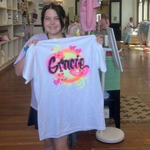 May include: White t-shirt with the name "Gracie" in a pink and black airbrushed design, surrounded by hearts and a yellow and orange gradient. The shirt is held up in a store setting.