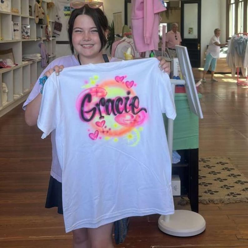 May include: White t-shirt with the name "Gracie" in a pink and black airbrushed design, surrounded by hearts and a yellow and orange gradient. The shirt is held up in a store setting.