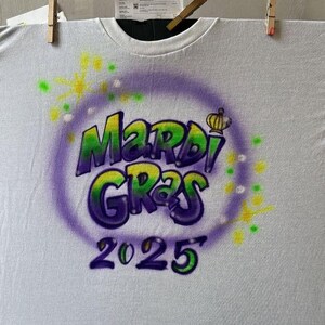 May include: White t-shirt with a purple, green, and yellow airbrushed design. The design features the words "Mardi Gras 2025" in a bubble letter style, surrounded by a purple circle and starburst accents. The shirt is hanging with clothespins.
