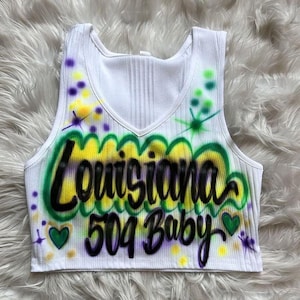 May include: White ribbed crop top with a V-neck. The top has airbrushed text reading "Louisiana 504 Baby" in black, yellow, and green. It's decorated with purple and yellow paint splatters and green heart shapes.