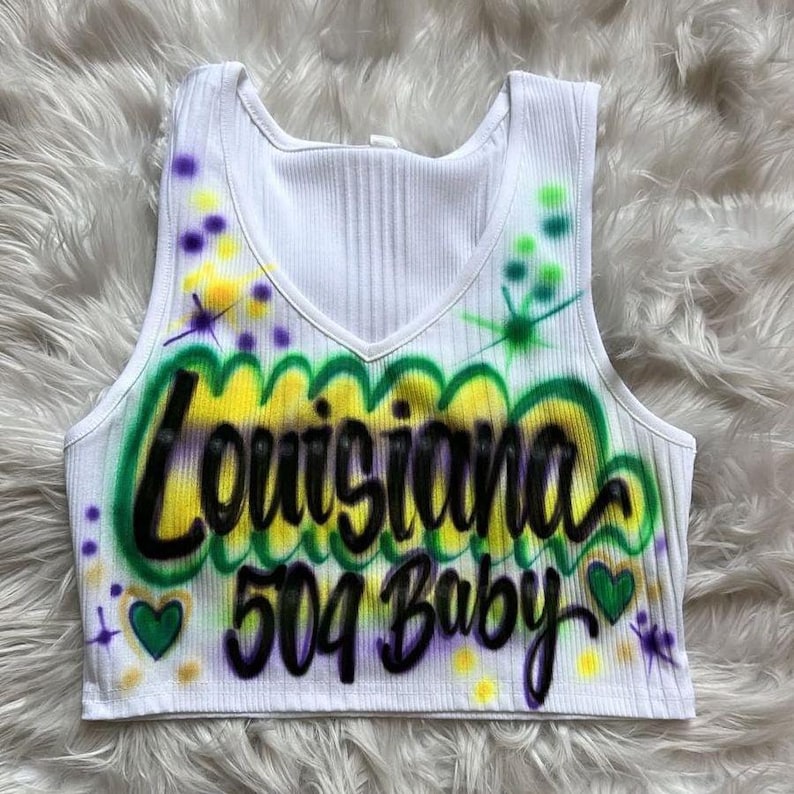 May include: White ribbed crop top with a V-neck. The top has airbrushed text reading "Louisiana 504 Baby" in black, yellow, and green. It's decorated with purple and yellow paint splatters and green heart shapes.