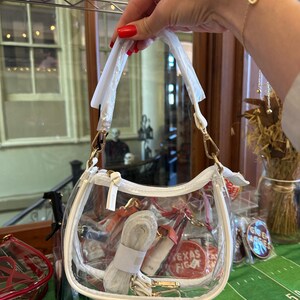 May include: A clear, white-trimmed handbag with a shoulder strap and gold-colored hardware. The bag contains a rolled-up strap and other items. The words "TEXAS FIGHT" are visible on a red circular item inside the bag.