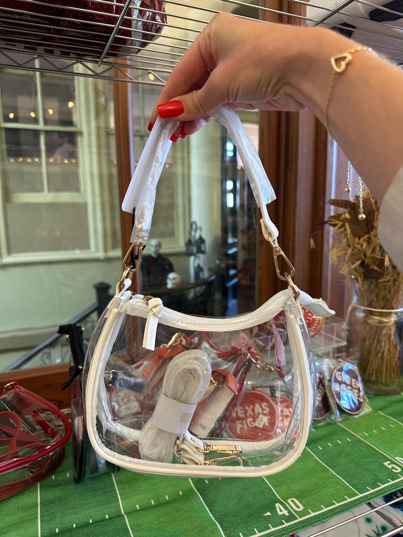 May include: A clear, white-trimmed handbag with a shoulder strap and gold-colored hardware. The bag contains a rolled-up strap and other items. The words "TEXAS FIGHT" are visible on a red circular item inside the bag.