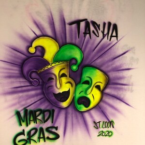 May include: White t-shirt with an airbrushed Mardi Gras design. The design features two theatrical masks, one smiling and one frowning, in yellow and green, with a purple and yellow jester hat. The words "TASHA", "MARDI GRAS", and "ST. LOUIS 2020" are also included.