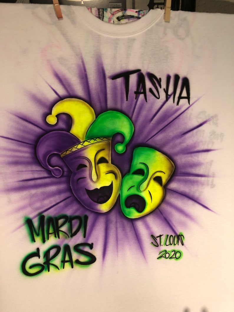 May include: White t-shirt with an airbrushed Mardi Gras design. The design features two theatrical masks, one smiling and one frowning, in yellow and green, with a purple and yellow jester hat. The words "TASHA", "MARDI GRAS", and "ST. LOUIS 2020" are also included.