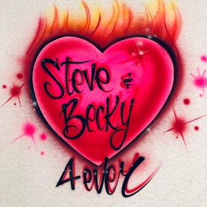 May include: A white t-shirt with a pink heart on fire. The heart has the words "Steve & Becky Forever" written on it.