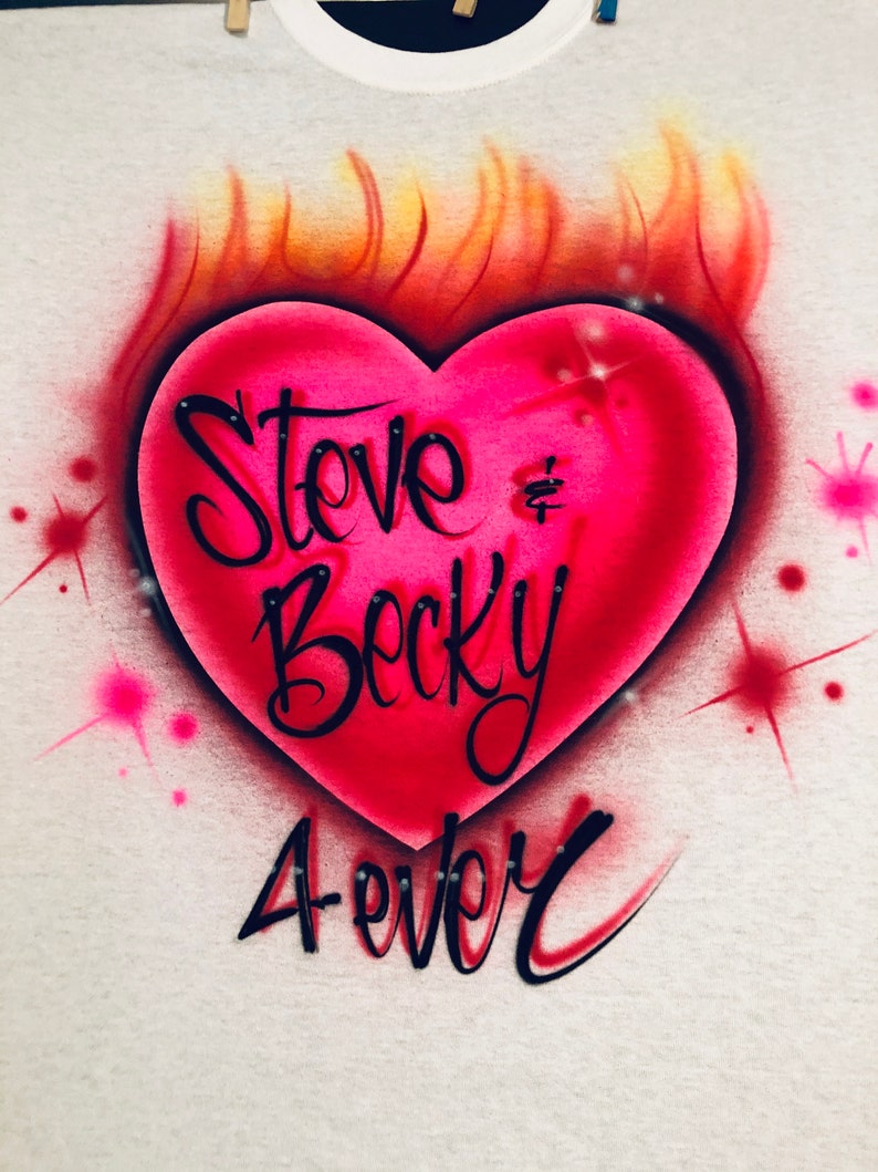 May include: A white t-shirt with a pink heart on fire. The heart has the words "Steve & Becky Forever" written on it.