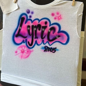 May include: White t-shirt with airbrushed design. The word "Lyric" is in a bubble letter style, with pink and blue gradients. The number "2005" is below the word. Pink and blue starbursts and dots are also airbrushed on the shirt.