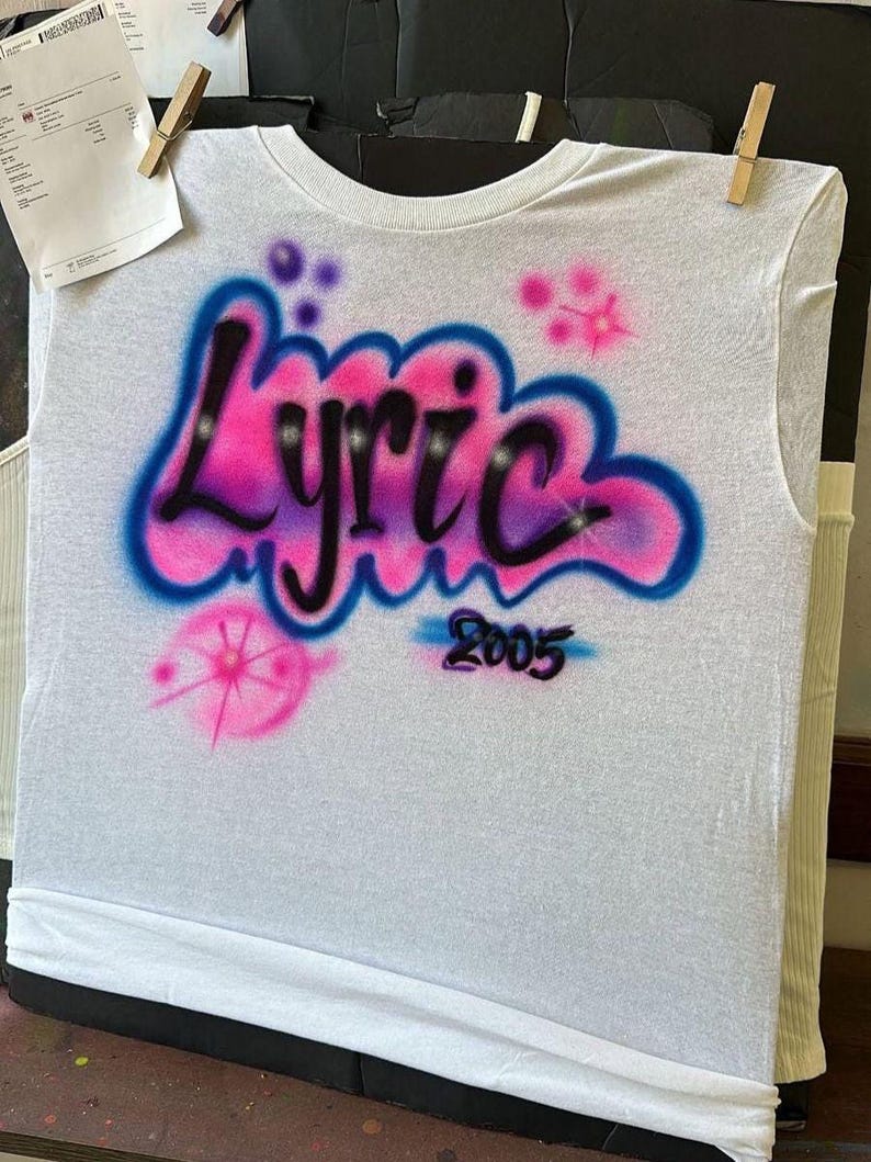 May include: White t-shirt with airbrushed design. The word "Lyric" is in a bubble letter style, with pink and blue gradients. The number "2005" is below the word. Pink and blue starbursts and dots are also airbrushed on the shirt.