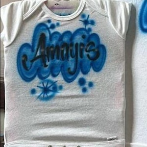 May include: White baby onesie with short sleeves. The onesie features the name "Amayis" in black lettering, surrounded by a blue cloud design. Additional blue starburst and dot accents are also present on the front.