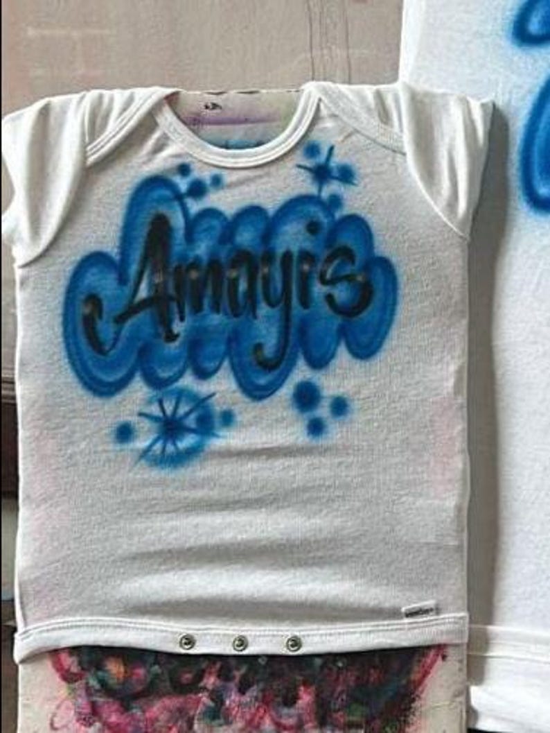 May include: White baby onesie with short sleeves. The onesie features the name "Amayis" in black lettering, surrounded by a blue cloud design. Additional blue starburst and dot accents are also present on the front.