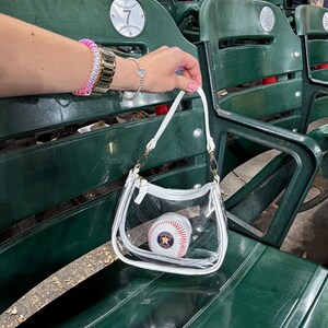 May include: A clear, white-trimmed handbag with a baseball inside, held in front of green stadium seating. The baseball has red stitching and a Houston Astros logo. The bag has a gold chain strap.