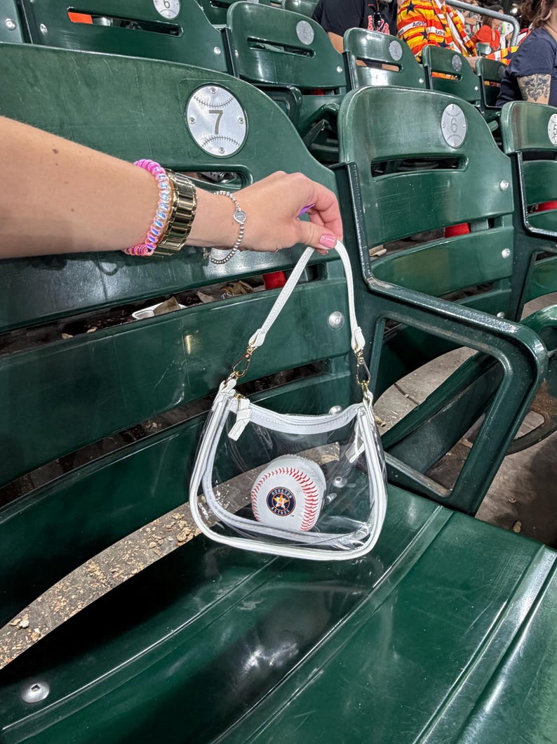 May include: A clear, white-trimmed handbag with a baseball inside, held in front of green stadium seating. The baseball has red stitching and a Houston Astros logo. The bag has a gold chain strap.