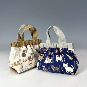 May include: Two small tote bags with handles. One bag is off-white with a dog pattern and brown handles. The other is navy blue with a dog pattern and gray handles. Both bags have a gathered top.