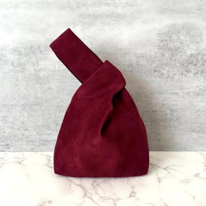 May include: A burgundy suede knot bag with a black trim. The bag is made of soft, supple suede and has a unique knot design.