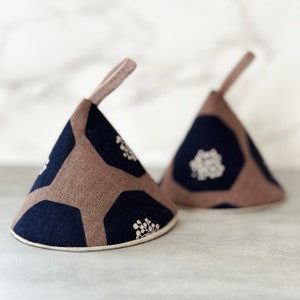 May include: Two brown and blue fabric pot holders with a white floral pattern. The pot holders are shaped like cones and have a loop handle.
