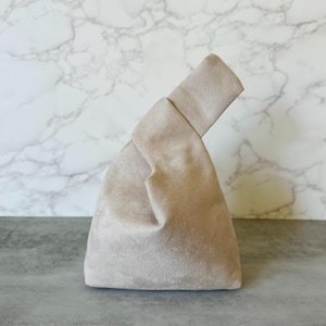 May include: A beige handbag with a knotted handle. The bag has a triangular shape and is made of a soft, suede-like material. The background is a marble pattern.