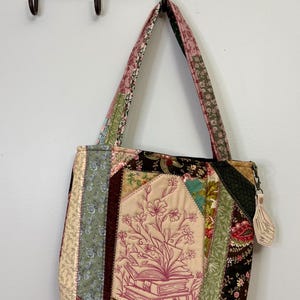 May include: A patchwork tote bag with a floral and book design. The bag features a variety of fabric patterns in shades of beige, green, pink, and black. The bag is hanging on a decorative metal hook.
