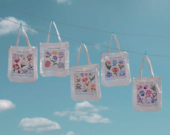 Pode incluir: Cinco sacolas brancas com ilustrações florais penduradas em um varal contra um céu azul. Cada sacola apresenta um design botânico diferente e o texto "FLORA OF LIVUE", "FLORA OF SUMERU", "FLORA OF MONDSTADT", "FLORA OF FONTAINE" e "FLORA OF INAZUMA".