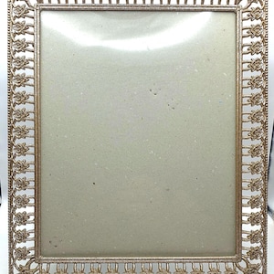 May include: A gold-toned metal picture frame with a decorative floral design. The frame is rectangular and has a simple, elegant design.