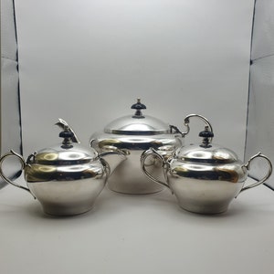 May include: A silver tea set comprising a teapot, creamer and sugar bowl. Each piece features a black knob on the lid.