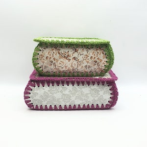 May include: Two crocheted boxes, one with a floral pattern and a green crocheted trim, and the other with a white lace pattern and a purple crocheted trim.