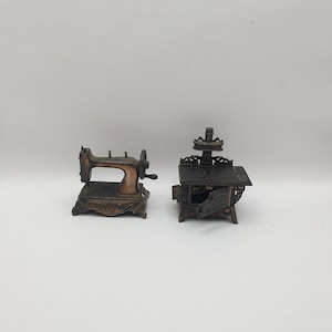 May include: Two vintage metal pencil sharpeners, one shaped like a sewing machine and the other shaped like a stove. Both are in a worn, antique bronze finish.