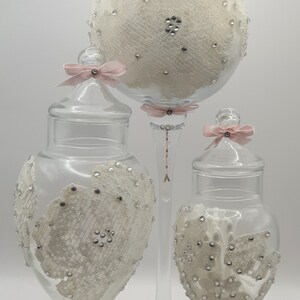 May include: Three glass jars with decorative lace and rhinestone accents. The jars have pink ribbon bows on the lids and are on clear glass pedestals.