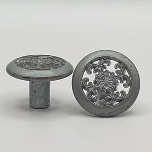 May include: Two silver metal cabinet knobs with a decorative, ornate, circular design. The knobs have a raised, circular base and a short, cylindrical stem.