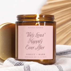 May include: A lit candle in a brown glass jar with a gold lid. The label reads "They Lived Happily Ever After" with the names Ashley + Mark and the engagement date. The candle sits on a white and black patterned cloth.