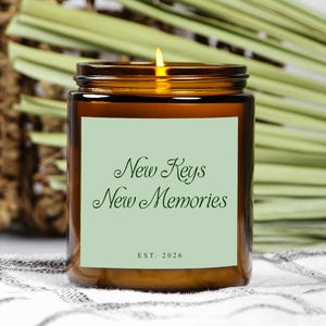May include: A lit brown glass candle jar with a light green label. The label reads "New Keys New Memories" with "EST. 2026" below. The candle sits on a white and gray patterned surface with a woven basket and green leaves in the background.