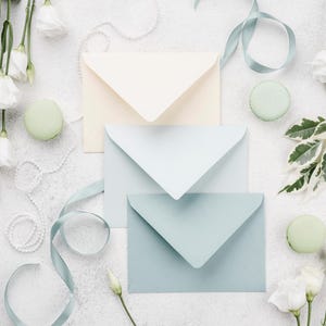 May include: Three stacked envelopes in cream, light blue, and sage green, accompanied by white flowers, green leaves, light green macarons, a string of white pearls, and a light blue ribbon, arranged in a flat lay composition.