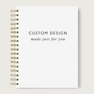 May include: A white spiral-bound notebook with gold-colored metal rings. The cover features the text "CUSTOM DESIGN made just for you" in a minimalist black font. The notebook is set against a neutral background.