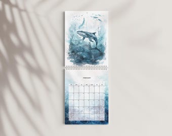 May include: A wall calendar for the month of February featuring a watercolor illustration of a whale swimming in deep blue ocean waters with seaweed. The calendar has a white grid for dates and is bound with a silver metal spiral at the center.