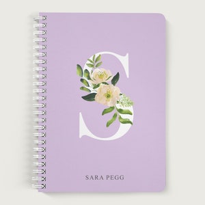 May include: A lavender notebook with a white letter S decorated with white flowers and green leaves. The name "SARA PEGG" is printed below the letter.