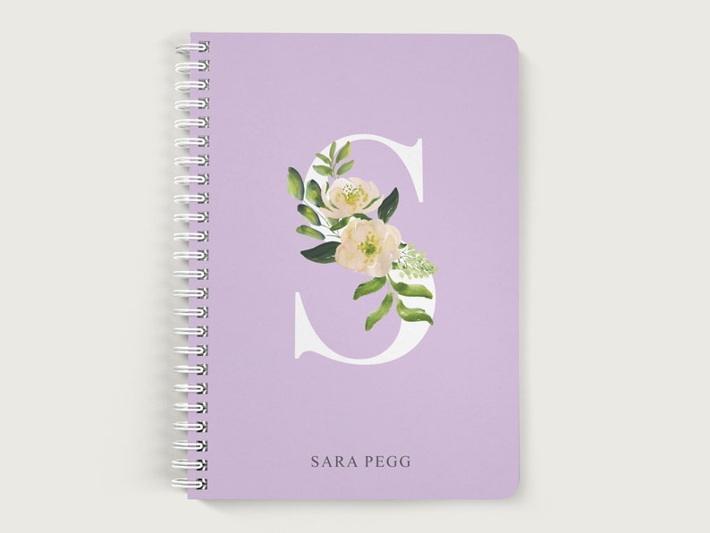 May include: A lavender notebook with a white letter S decorated with white flowers and green leaves. The name "SARA PEGG" is printed below the letter.