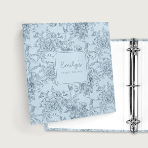 May include: A light blue binder with a floral pattern and the text "Emily's Family Recipes." The binder is open, revealing a three-ring mechanism and white pages. The floral design features flowers and leaves.